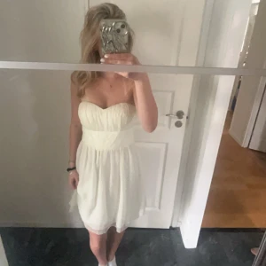 Vit axelbandslös klänning - Säljer en söt vit axelbandslös klänning med hjärtformad byst och markerad midja. Klänningen har lätt rynkat tyg och en luftig kjol som slutar ovanför knäna. Perfekt för sommarens alla tillfällen!