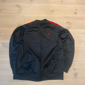 Svart track jacket med röda ränder - Snygg svart tröja  med röda ränder längs ärmarna och liten röd logga på bröstet. Jackan har dragkedja framtill, ribbade muddar och två ränder på ärmarna. Skicka ett meddelande för frågor, storlek, mått osv. 