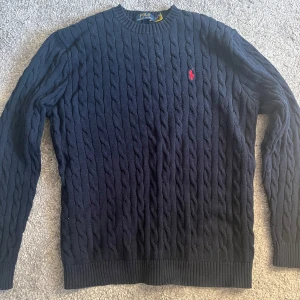 Mörkblå kabelstickad tröja från Ralph Lauren - Säljer en klassisk mörkblå kabelstickad tröja från Ralph Lauren med röd broderad logga på bröstet. Tröjan har rund halsringning och långa ärmar. Perfekt för en stilren och tidlös look. Priset är ej hugget i sten!