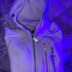 Vit jacka med huva från Sail Racing - Säljer en vit jacka från Sail Racing med huva och dragkedjor framtill. Jackan har en bröstficka med dragkedja och logga på bröstet. Perfekt för dig som gillar sportig stil och vill ha något snyggt och funktionellt.