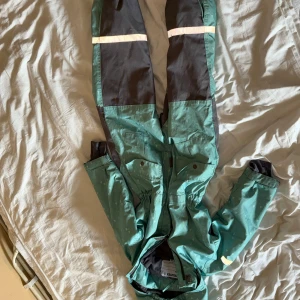 Grön och svart täckbyxdress från Snowtex - Grön täckbyxdress med svarta förstärkningar på knän och bak, samt reflexdetaljer. Perfekt för kalla dagar med elastisk midja och dragkedja framtill. Märket Snowtex syns på insidan. Snygg och praktisk overall för vinteraktiviteter.