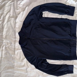 Mörkblå bomberjacka från COS - Snygg mörkblå bomberjacka från COS med dragkedja framtill och ribbade muddar vid ärmslut, krage och nederkant. Jackan har en stilren design och är tillverkad i ett slätt syntetmaterial. Perfekt för dig som gillar minimalistisk stil.