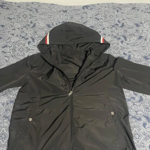 Svart vindjacka från Moncler med huva - Säljer en svart vindjacka från Moncler med huva och dragkedja. Jackan har Moncler-logga på ärmen och vita samt röda detaljer på huvan. Perfekt för blåsiga dagar och snygg streetstil. Byten är även intressant 