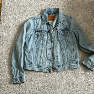 Ljusblå jeansjacka från Levi's - Säljer en klassisk ljusblå jeansjacka från Levi's i storlek S. Jackan har knappar framtill, två bröstfickor med lock och knapp samt två sidofickor. Perfekt för dig som gillar en tidlös och avslappnad stil.
