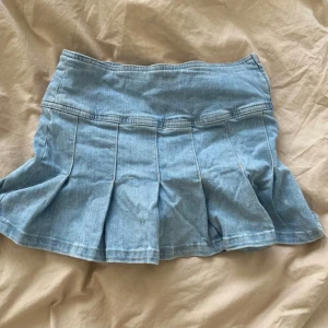 Ljusblå jeanskjol med dragkedja - Säljer en kort lågmidjad ljusblå jeanskjol med veckad design och dragkedja i sidan. Kjolen har en markerad midja och klassisk jeanssöm. Perfekt för dig som gillar en trendig och avslappnad stil.