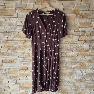 Brun prickig klänning från Lindex - Säljer en brun klänning med vita prickar från Lindex. Klänningen har v-ringning, knappar framtill och korta ärmar. Den har en avslappnad passform och är tillverkad i ett mjukt material som känns skönt mot huden.