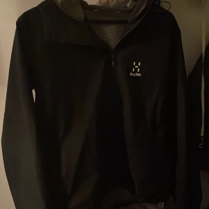 Svart vindjacka från Haglöfs - Säljer en svart vindjacka från Haglöfs med halv dragkedja framtill och hög krage. Jackan har en sportig look och är perfekt för utomhusaktiviteter. Klassisk logga på bröstet och lätt material som andas.