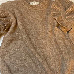 Beige/Brun stickad tröja med volang från H&M - Säljer en brun stickad tröja från H&M med långa ärmar och volangdetaljer på axlarna. Tröjan har rund halsringning och är mjuk och skön, perfekt för kyligare dagar.