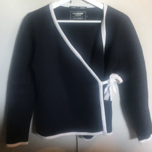 Svart omlottkofta med vit kant från Holebrook - Säljer en svart omlottkofta från Holebrook med vita kanter och knytband i sidan. Koftan har lång ärm och är perfekt för lager-på-lager. Enkel och stilren design som passar till mycket.