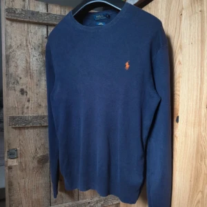 Blå tröja från Polo Ralph Lauren - Säljer en klassisk blå tröja från Polo Ralph Lauren i slim fit-modell. Tröjan är tillverkad i mjuk pima bomull och har lång ärm samt den ikoniska broderade loggan i orange på bröstet.