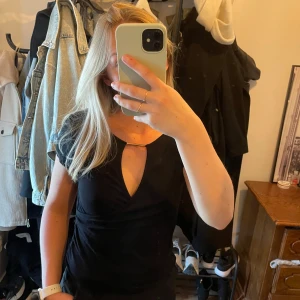 Svart topp med cut-out från Happy Holly - Säljer en svart topp från Happy Holly med korta ärmar och snygg cut-out detalj framtill. Toppen har en lätt drapering på ena sidan och är tillverkad i ett mjukt material som sitter bekvämt. Perfekt för att lyfta din outfit!