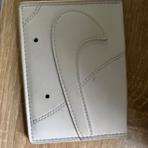 Vit plånbok från Nike - Säljer en stilren vit plånbok från Nike i syntetmaterial. Plånboken har det klassiska Nike-loggan och swoosh-detaljen i relief på framsidan. Enkel och smidig design med plats för kort och sedlar.