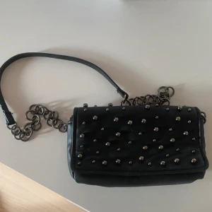 Svart axelväska med nitar och kedja - Svart axelremsväska i skinnimitation med silverkulor. Från Zara. Höjd: 10,5 cm, bredd: 20 cm, djup: 5 cm.