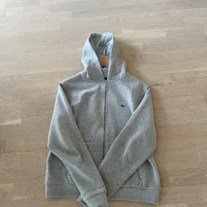 Grå hoodie från Lacoste - Säljer en klassisk grå hoodie från Lacoste med dragkedja och huva. Tröjan har det ikoniska krokodilmärket broderat på bröstet och är tillverkad i mjuk bomull. Perfekt för en avslappnad stil.