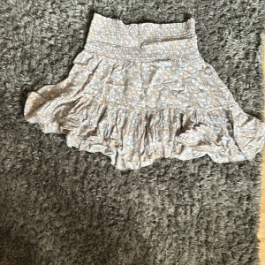 Beige blommig volangkjol från 157 - Supersöt beige kortkjol från 157 med vitt blommönster och volangdetaljer. Kjolen har smock i midjan för en bekväm passform och är perfekt för varma dagar. Lätt och luftig känsla.