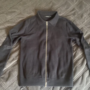 Svart Matinique tröja med dragkjedja - Säljer en svart cardigan jacka från Matinique med hel dragkedja framtill och klassisk krage. Tröjan är långärmad och har ribbade muddar vid ärmslut och nederkant. Perfekt för lager-på-lager och enkel att matcha. Änden på ena Armen har gått upp.