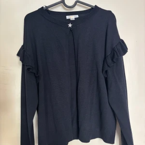 Mörkblå stickad tröja med volangdetaljer - Mörkblå stickad tröja från Gina Tricot med långa ärmar och volangdetaljer längs axlarna. Tröjan har en liten silverfärgad stjärndetalj vid halsringningen och ribbade muddar vid ärmslut och nederkant.