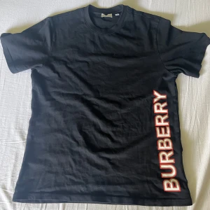 Svart Burberry t-shirt - Säljer en svart t-shirt från Burberry med stor vit och röd logga längs sidan. Storleken är XS men passar både XS och S. Jag säljer den eftersom den inte kommer till användning längre. För mer info hör av er. Priset är diskuterbart