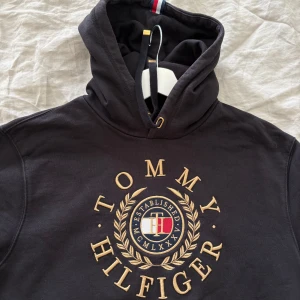 Svart hoodie från Tommy Hilfiger - Svart hoodie med stor broderad Tommy Hilfiger-logga och gulddetaljer framtill. Klassisk huva med snörning och magficka. Perfekt för en avslappnad stil. Passar både storlek M och L