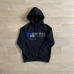 Svart Trapstar Tracksuit  - Svart hoodie från Trapstar i storlek S med huva och snörning. Framsidan har ett stort Trapstar-tryck i blått, grått och vitt. Klassisk känguruficka och ribbade muddar. Perfekt för dig som gillar streetwear och vill sticka ut med en snygg logga.