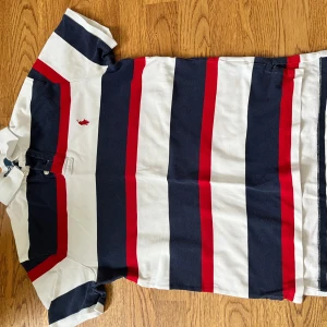 Randig pikétröja från Polo Ralph Lauren - Snygg pikétröja från Polo Ralph Lauren med breda marinblå, vita och röda ränder. Klassisk krage med knappar och broderad röd logga på bröstet. Tillverkad i mjuk bomull, perfekt för en sportig och stilren look. Inga skador eller smuts
