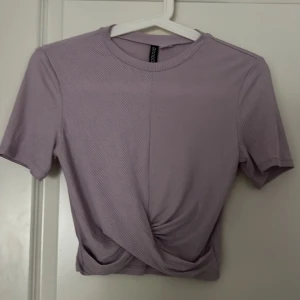 Ljuslila ribbad croppad topp - Säljer en ljuslila ribbad croppad topp med korta ärmar och twistad detalj framtill. Perfekt för vår och sommar, enkel att matcha med både jeans och kjol. Storlek S
