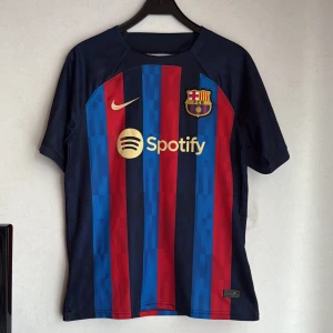FC Barcelona hemmatröja 2022/23 - Säljer en officiell FC Barcelona hemmatröja från säsongen 2022/23. Tröjan är kortärmad med klassiska blå och röda vertikala ränder, Nike-logga och Spotify som sponsor på bröstet. Broderat klubbmärke och UNHCR-tryck på ryggen. Tillverkad i lätt och ventilerande Dri-FIT-material. Tröjan är köpt från Nike, så den är 100% äkta 