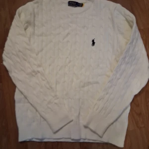 Vit kabelstickad tröja från Polo Ralph Lauren - Kabelstickad vit tröja från Polo Ralph Lauren med klassisk rund halsringning och broderad logga på bröstet. Tröjan har lång ärm och ribbade muddar vid ärmslut och nederkant. Perfekt för en stilren och tidlös look.