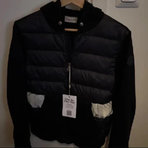 Svart pufferjacka från Moncler - Svart pufferjacka från Moncler med quiltad front och vita detaljer på fickorna. Jackan har dragkedja framtill, huva och långa ärmar. Perfekt för dig som vill ha en stilren och sportig look. Har äkthetsbevis och allting. 