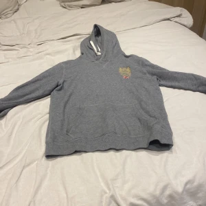 Grå hoodie från Morris med brodyr - Snygg grå hoodie från Morris med guldfärgad brodyr och brittisk flagga på bröstet. Klassisk känguruficka framtill, huva med vit dragsko och mjukt bomullsmaterial. Perfekt för dig som gillar stilrena och bekväma plagg med en touch av preppy.