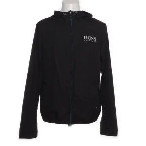 Svart vindjacka från Hugo Boss - Svart vindjacka från Hugo Boss med vit logga på bröstet och huvan. Jackan har dragkedja framtill och huva med snörning. Perfekt för dig som vill ha en stilren och sportig look.