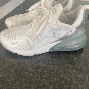 Nike Air Max 270 vita sneakers - Nike Air Max 270 sneakers i vitt med silvriga detaljer och stor synlig luftbubbla i hälen. Ovandel i mesh för bra andningsförmåga, snörning och klassisk Nike-logga på sidan. Perfekta för dig som gillar sportig och modern stil. Använda 2-3 gånger. 