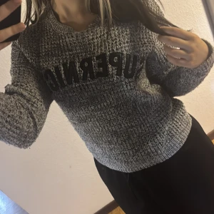 Grå stickad tröja med stjärnor - Säljer en gråmelerad stickad tröja från Supernice med svart text framtill och coola svarta stjärnor på ena ärmen. Tröjan har en loose passform och långa ärmar, perfekt för dig som gillar en avslappnad stil.