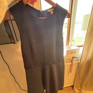 Svart ärmlös kortklänning med spets - Snygg svart kortklänning med ärmlös design och rund halsringning. Klänningen har dekorativ spetsdetalj i midjan som ger en extra touch. Perfekt för dig som gillar stilrena och klassiska plagg.