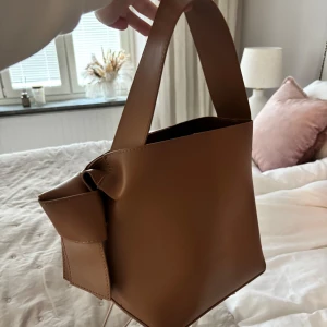 Brun bucket handväska med rosett - Snygg beige bucket handväska i slätt skinnimitation med bred axelrem och dekorativ rosett på sidan. Väskan har en innerficka med dragkedja och stilren, minimalistisk design som passar till många outfits.