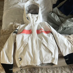 Vit vindjacka Helly Hansen XS - Säljer en vit vindjacka från Helly Hansen i storlek XS. Jackan har huva, svarta detaljer på ärmslut och nederkant, samt en rosa rand över bröstet. Innerkragen är fodrad med svart fleece och jackan har dragkedja framtill och praktiska fickor.