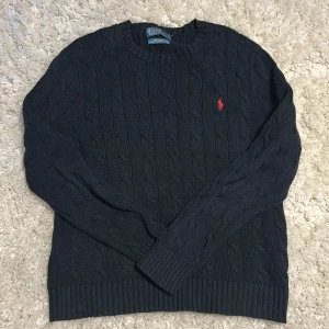 Mörkblå kabelstickad tröja från Polo Ralph Lauren - Kabelstickad mörkblå tröja från Polo Ralph Lauren i 100% bomull. Klassisk modell med rund halsringning och röd broderad logga på bröstet. Ribbstickade muddar vid ärmslut och nederkant. Perfekt för en stilren look.