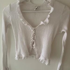 Vit ribbad tröja med volangdetaljer - Säljer en söt vit ribbad tröja med volangkant längs halsringning, knappslå och nederkant. Tröjan har långa ärmar och är croppad i modellen. 