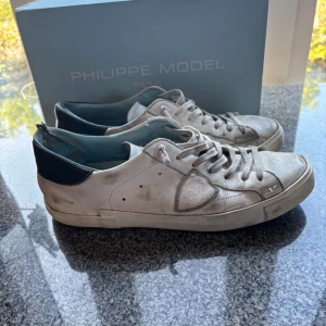 Philippe Model skor - Säljer ett par vita Philippe Model skor med svarta detaljer på hälen och klassisk snörning. Skorna har en rund tå och platt sula, tillverkade i skinn med en stilren och tidlös design. Perfekta för dig som gillar en clean look. Dom är dock ganska slitna inuti i hälen men är inget som syns på utsidan. Dom är storlek 43 men pågrund av att hälen är ganska sliten så passar dom en storlek större ungefär. Boxen tillhör. Priset är inte hugget i sten!