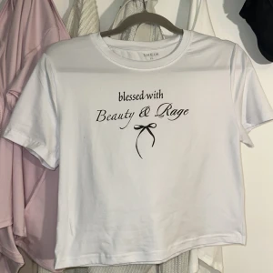 Vit croppad topp från SHEIN - Superfin vit croppad topp från SHEIN i nyskick, endast testad en gång! 🖤 Har texten “blessed with Beauty & Rage” och en söt rosett framtill. Storlek XL men stretchig, så den passar allt från XS till XXL beroende på passform, från loose till tight crop.