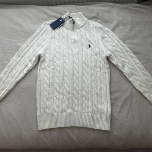 Vit kabelstickad tröja Polo Ralph Lauren - Vit kabelstickad tröja från Polo Ralph Lauren med halv dragkedja och hög krage. Klassisk broderad logga på bröstet. Tröjan är långärmad och har ribbade muddar och nederkant. Perfekt för lager-på-lager under kyliga dagar.