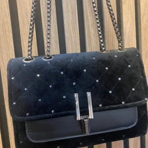 Svart quiltad axelväska med kedjerem - Snygg svart axelväska med quiltat mönster och små glittriga detaljer. Väskan har en silverfärgad kedjerem och ett stilrent spänne framtill. Perfekt för att lyfta din outfit med en elegant touch.
