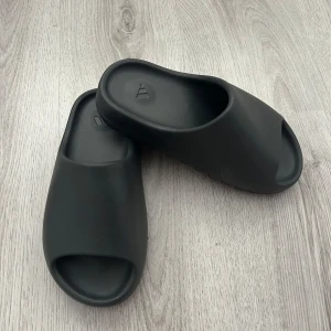 Svarta Adidas Yeezy Slides  - Helt nya Svarta Adidas Yeezy Slides med öppen tå och kraftig, räfflad sula. Tillverkade i syntetmaterial och har en minimalistisk design med Adidas-logga i fotbädden. Perfekta för sommaren och enkel att glida i.