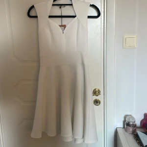 Vit ärmlös kortklänning med v-ringning - Superfin vit kortklänning med v-ringad hals och breda axelpartier. Klänningen är ärmlös och har en markerad midja med utsvängd kjol. Köpt från Nelly men aldrig kommit till användning. Perfekt inför sommaren!😍