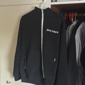 Palm Angels svart track jacket - Svart track jacket från Palm Angels med vita ränder längs ärmarna och vit dragkedja framtill. Jackan har logga på bröstet, två fickor med dragkedja och är gjord i bomull och polyester. Perfekt för en sportig och trendig look.