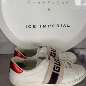 Gucci vita sneakers med röd detalj - Snygga vita sneakers från Gucci med breda elastiska band över foten med klassiskt Gucci-tryck i rött och blått. Skorna har röda metallic-detaljer på hälen och är tillverkade i skinn. Sulan har ett unikt Gucci-mönster undertill. Skick 9/10