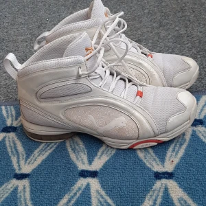 Vita höga sneakers från Puma - Snygga vita höga sneakers från Puma med coola mönsterdetaljer på sidorna och orange logga. Skorna har snörning, meshpaneler för extra andning och inslag av syntet och skinn. Sulan har röda detaljer och Puma-logga undertill. Perfekta för dig som gillar sportig stil.