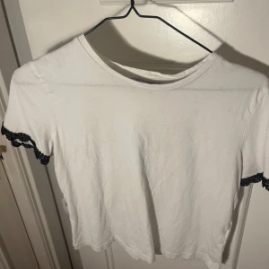 Vit topp med svarta spetsdetaljer - Säljer en söt vit topp med korta ärmar och svarta spetsdetaljer vid ärmsluten. T-shirten är simpel med lite detalj🩷