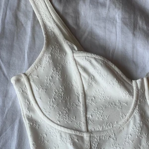 Vit korsettbody med broderade detaljer - Supersnygg vit korsettinspirerad body med broderade blommönster och breda axelband. Figurnära passform med markerade kupor och knäppning i grenen. Perfekt att styla med jeans eller kjol för en trendig look.