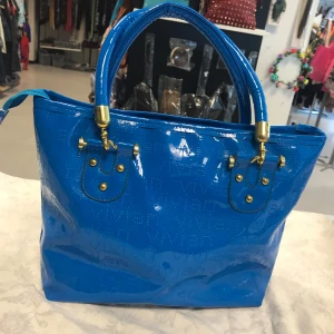 Blå glansig handväska med gulddetaljer - Snygg blå handväska i glansigt syntetmaterial med två stadiga handtag och guldfärgade metalldetaljer. Väskan har en dragkedja på baksidan och stilren design, perfekt för att lyfta din outfit. Oanvänd 